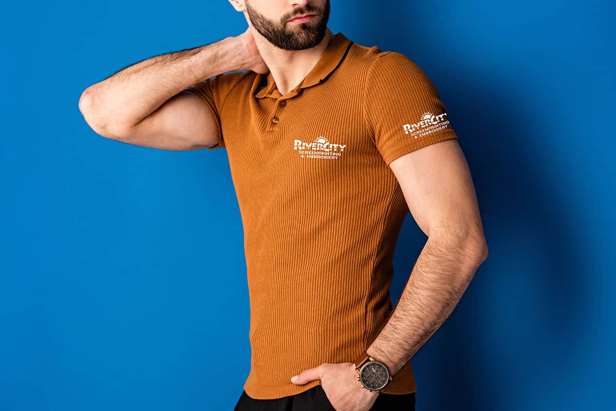 Man Wearing Polo A man in a brown ribbed polo shirt stands against a blue background, with one hand on his neck and the other resting at his side. He is wearing a watch on his left wrist, and the shirt features white text on the left chest and right sleeve.